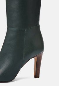 Bottine en cuir vert foncé avec un bout pointu et un talon bloc beige moyen, présentée sur un fond blanc.