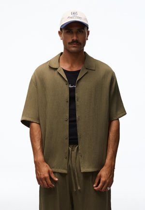 Mann mit Schnurrbart, der eine beige Baseballkappe trägt, ein lockeres olivgrünes kurzärmeliges Hemd mit Knöpfen über einem schwarzen Shirt und passende olivgrüne Hosen.