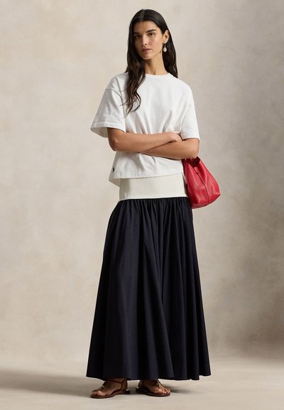 Λευκό κοντό t-shirt, ελαφρύ ύφασμα· μαύρη μακριά φούστα με πιετά στη μέση· κόκκινη δερμάτινη τσάντα· καφέ επίπεδα σανδάλια· ουδέτερο φόντο.