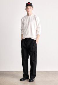 Λευκή μπλούζα με μικρό κόκκινο λογότυπο, μαύρο παντελόνι cargo με πλάγιες τσέπες, μαύρη ζώνη και μαύρα παπούτσια, όλα σε μια ουδέτερη φόντο.