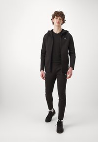 Melnā sportiskā treniņtērps ar kapuci un pieguļošām biksēm, izgatavots no gludas auduma, ar logotipa akcentu pie krūšu un augšstilba.