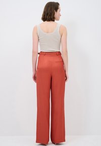 Rust-colored, high-waisted trousers in a loose fit with a straight leg, featuring a back pocket and smooth fabric texture.