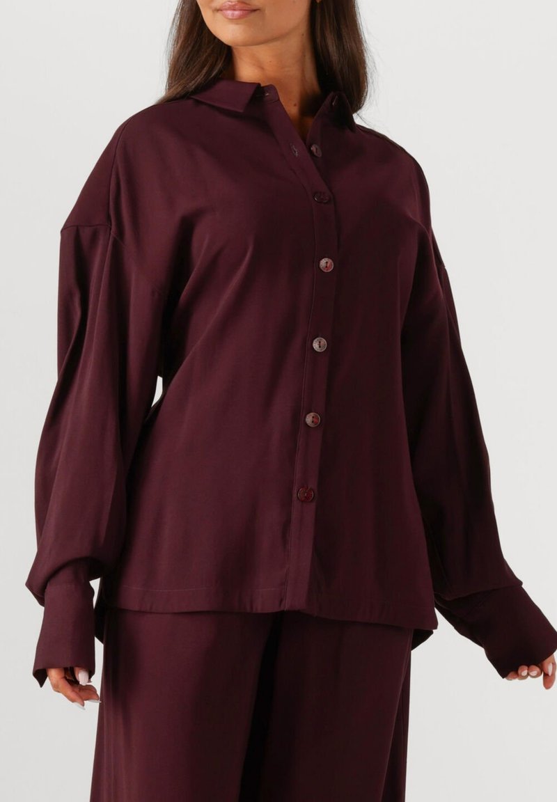 Burgundy button-up shirt with long sleeves and a classic collar. Smooth fabric with a relaxed fit, featuring round buttons down the front.