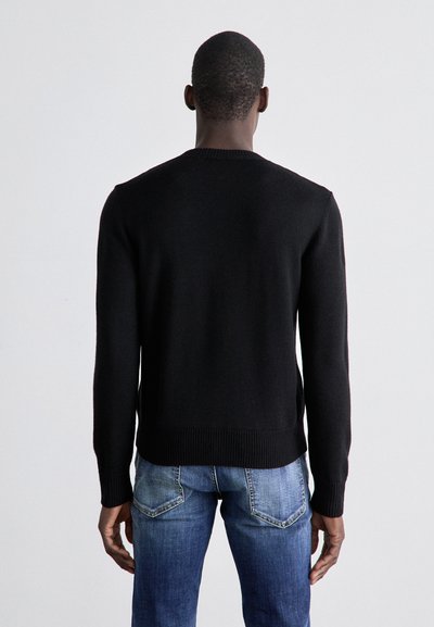 Sort strikket sweater med rund hals, featuring en ribbet kant og manchetter, båret over lyseblå jeans. Teksturen ser blød og tætsiddende ud.