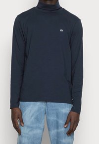 Mörkblå polotröja i mjukt tyg med långa ärmar, prydd med en liten vit logotyp på bröstet, kombinerad med ljusblåjeans.