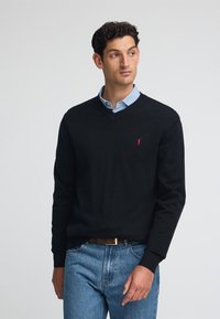 Maglione nero a V in materiale a maglia, con un piccolo logo rosso sul petto, abbinato a una camicia colletto azzurro e jeans blu.