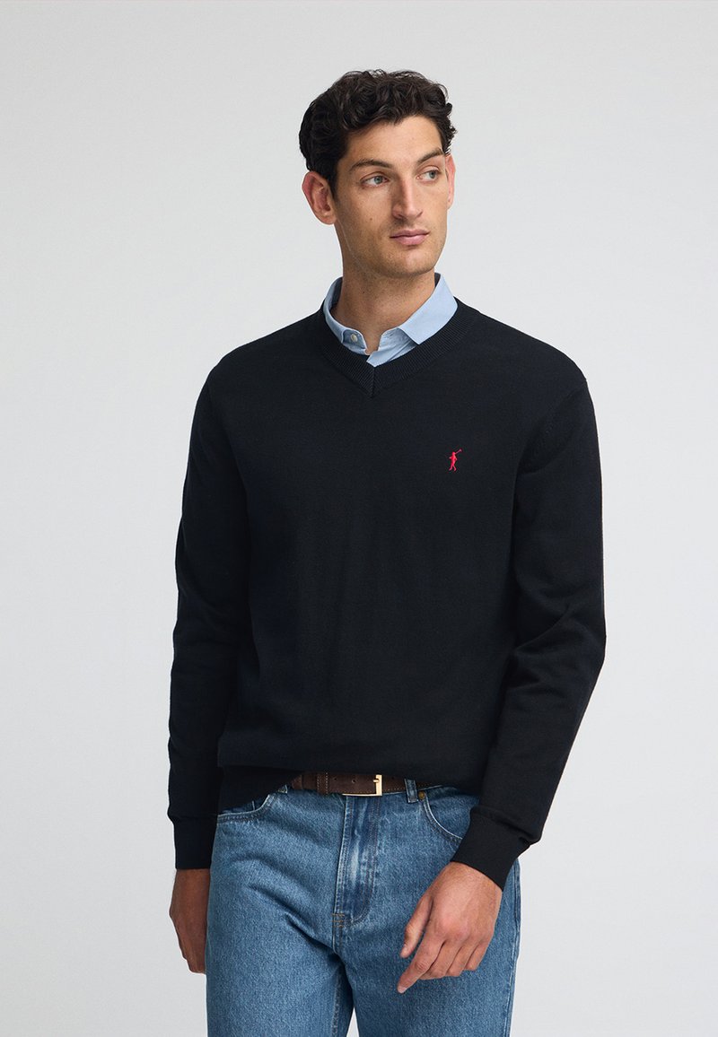 Maglione nero a V in materiale a maglia, con un piccolo logo rosso sul petto, abbinato a una camicia colletto azzurro e jeans blu.