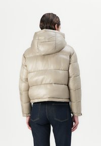 Pufferjack in lichtbeige met een glanzende afwerking, met horizontale stiksels, een capuchon en elastische manchetten. Gecombineerd met donkerblauwe jeans.