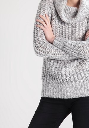 Grijze gebreide trui met een colkraag, een gestructureerd patroon en een geribde zoom. Gecombineerd met zwarte spijkerbroek.