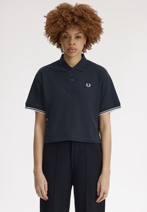 Mujer con cabello rizado que lleva una camiseta tipo polo azul marino oscuro con un logo de corona de laurel y pantalones plisados negros, de pie frente a un fondo blanco.