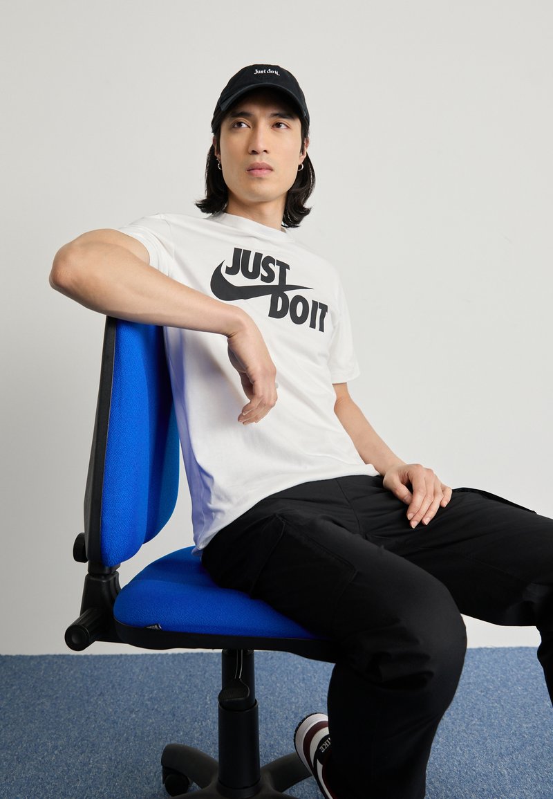 Hvid t-shirt med "Just Do It" i sort, sort kasket, sorte bukser, siddende på en blå ergonomisk stol med tekstureret stof.