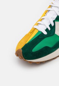 Avant de chaussure de sport avec daim vert, panneaux en tissu jaune et blanc, lacets blancs, et semelle en caoutchouc marron texturée.