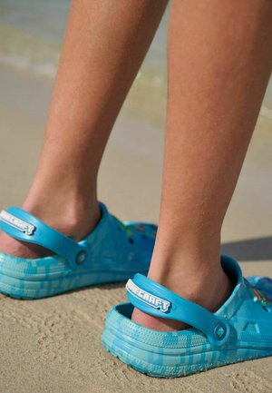 Blå lätta träskor med justerbara bakremmar, sulor med texturerat grepp och Minecraft-temade mönster; sedda bakifrån på en sandig strand.