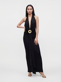Woman with long dark hair wearing a black halter-neck gown with a gold circular buckle at the waist and gold high-heeled sandals.