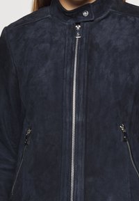 Námořnická modrá surová sukně s vysokým límcem, centrálním zipem a dvěma bočními zipy. Měkká textura s elegantním, minimalistickým designem.