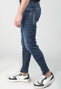Jeans de denim oscuros y ajustados con una textura suave, que presentan un bordado intrincado y bolsillos traseros, combinados con zapatillas negras y blancas.
