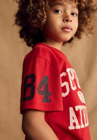 Rød bomulds t-shirt med hvid "SPORT ATH" tekst og en sort "84" på ærmet. Standard rund hals og korte ærmer.