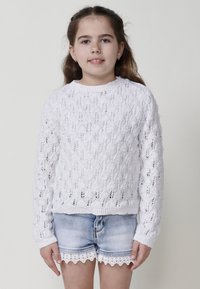 Pull en tricot blanc avec un motif en diamant texturé, associé à un short en denim bleu clair avec un détail de bord en dentelle.