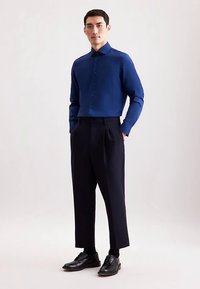 Donkerblauw overhemd met knopen en een scherpe kraag, gecombineerd met hoge zwarte broeken en zwarte leren schoenen, wat een op maat gemaakte uitstraling creëert.