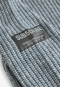 Graues gestricktes Gewebe mit einem strukturierten Muster. Enthält ein schwarzes Etikett mit weißem Text, auf dem "SUB5CRIBE LDN" steht, sowie weiteren Details darunter.