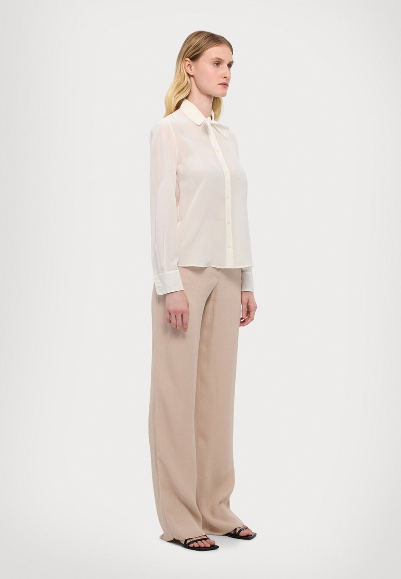 Transparente weiße Bluse mit Kragen, kombiniert mit beigefarbenen weiten Hosen und schwarzen Riemchensandalen, seitlich präsentiert.
