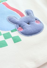 Tissu en coton avec un appliqué de lapin bleu aux joues roses, entouré de formes colorées dans des tons de bleu, vert et rouge.