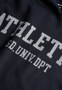 Marineblåt stof med stor hvidt trykt tekst "ATHLET" og mindre tekst "SD. UNIV. DPT" i fed, kollegiestil skrifttype.