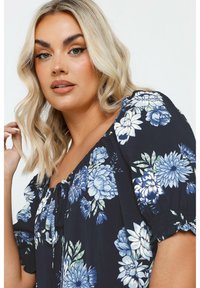 Blusa floral con fondo azul marino, adornada con flores azules y blancas, mangas fruncidas y escote con cordones. Tejido suave y ligero.