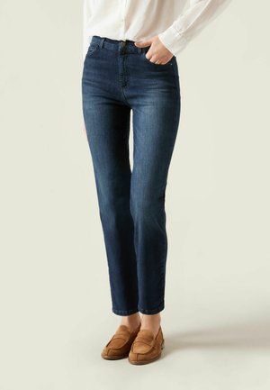 Jeans de mezclilla en un lavado oscuro con un corte recto, diseño de cinco bolsillos, cierre de botón y detalles de costura sutiles, acompañados de mocasines marrones.