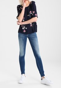 Top navy con ricami floreali, maniche corte, abbinato a jeans skinny blu chiaro e sneakers bianche. Design semplice, vestibilità casual.