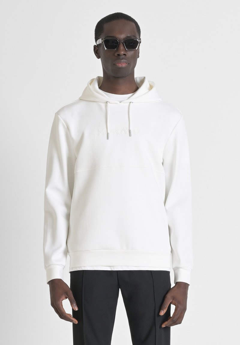 Witte hoodie van zacht materiaal, met een kangoeroezak, trekkoordcapuchon en subtiel reliëflogo. Gecombineerd met zwarte broek en zonnebril.