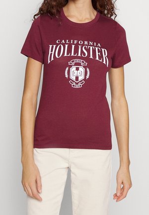 Bordo koszulka z krótkim rękawem z białym napisem „California Hollister Since 1999” i emblematem, noszona z jasnobeżowymi spodniami.