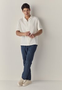 Camicia bianca a maniche corte con texture e colletto; abbinata a pantaloni in denim blu e sneakers beige. Il soggetto è appoggiato in modo casual a un muro.