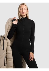 Zwarte geribbelde jumpsuit met een hoge kraag en rits, gecombineerd met een beige pufferjack. De stof heeft een gladde textuur en een getailleerd ontwerp.