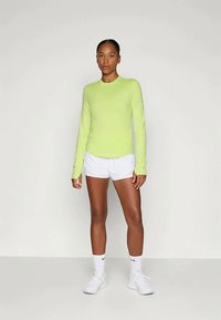 Donna in piedi che indossa una maglietta a maniche lunghe verde lime, pantaloni corti sportivi bianchi, calze bianche e scarpe da ginnastica bianche su uno sfondo chiaro e neutro.