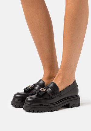 Mocassins en cuir noir avec une finition lisse, ornés de glands décoratifs et de quincaillerie dorée, reposant sur une semelle en caoutchouc noire épaisse.