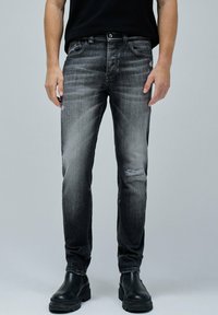 Jeans de mezclilla negra, corte slim con desgastes y ligeros desgarros en las rodillas. Combinados con botas negras hasta el tobillo, contra un fondo gris.