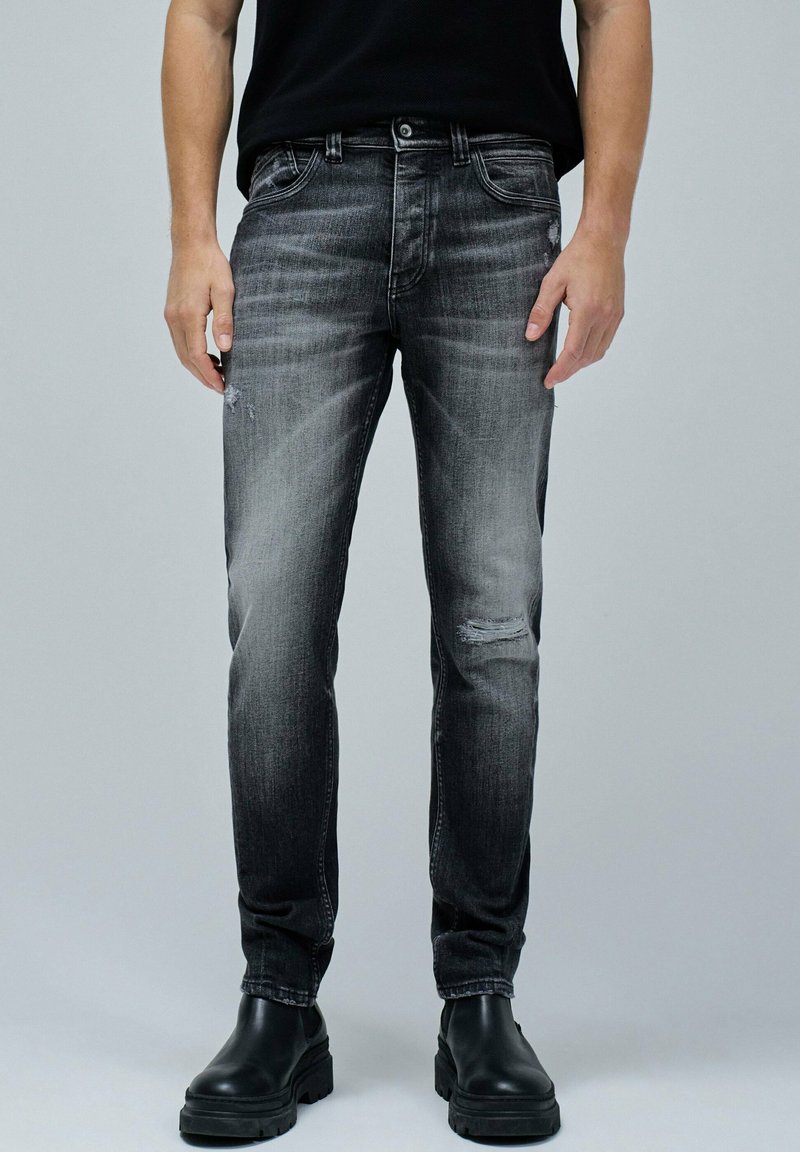 Jeans de mezclilla negra, corte slim con desgastes y ligeros desgarros en las rodillas. Combinados con botas negras hasta el tobillo, contra un fondo gris.