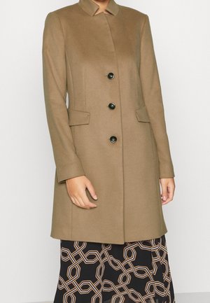 Kvinde iført en kamelfarvet frakke med sorte knapper over et sort nederdel med et beige geometrisk kædemønster.