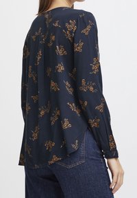 Langærmet bluse i marineblåt stof med blomsterprint i brunt og guld. Rundet bageste kant og plisseret design ved skuldrene.