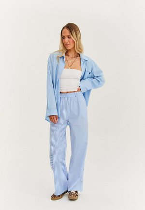 Žena v světle modré oversized košili, bílé trubkové topu, modro-bílých pruhovaných širokých kalhotách, botách s leopardím vzorem a s vrstvami náhrdelníků.