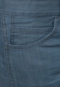 Tummansiniset farkut, joissa on edessä kaareva tasku, näkyvät saumat ja pehmeä, teksturoitu kangas.