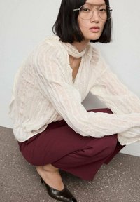 Blusa blanca con textura y mangas largas; pantalones burdeos oscuros; tacones negros de punta abierta. Gafas transparentes y detalles de diseño minimalista.