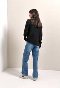 Pull en maille texturée noire, coupe ample, associé à un jean taille haute bleu et des baskets blanches avec un logo rouge. Fond neutre.