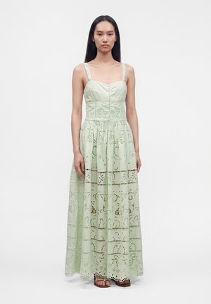Jeune femme aux longs cheveux noirs portant une robe longue sans manches vert pâle avec des motifs floraux découpés et des sandales marron, debout sur fond blanc.