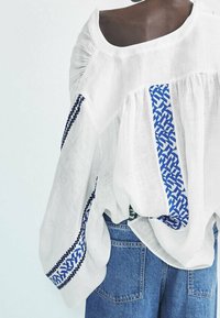 Witte linnen blouse met brede mouwen, versierd met blauwe geometrische borduursels aan de voorkant en achterkant. Draagt bij blauwe denim jeans.