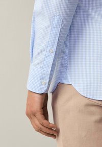Camicia a scacchi azzurro chiaro con polsini abbottonati, caratterizzata da un orlo curvo e bottoni bianchi a contrasto. Il materiale appare liscio e leggero.
