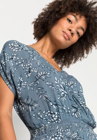 Bluza od blagog materijala s floralnim uzorkom u tirkiznoj boji, s V-izrezom, kratkim rukavima i strukiranjem. Materijal izgleda lagano i ima meku teksturu.