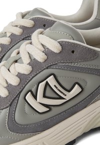 Pelēkie ādas un zamšādas sporta apavi ar krēmkrāsas šņorēm, ar teksturētu virsmu un kontrastējošu logo akcentu. Izstrādāti ar biezu zoli.