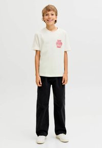 Crème-kleurig katoenen t-shirt met een rode vaasprint op de borst, gecombineerd met losse donkergekleurde broek en witte sneakers.